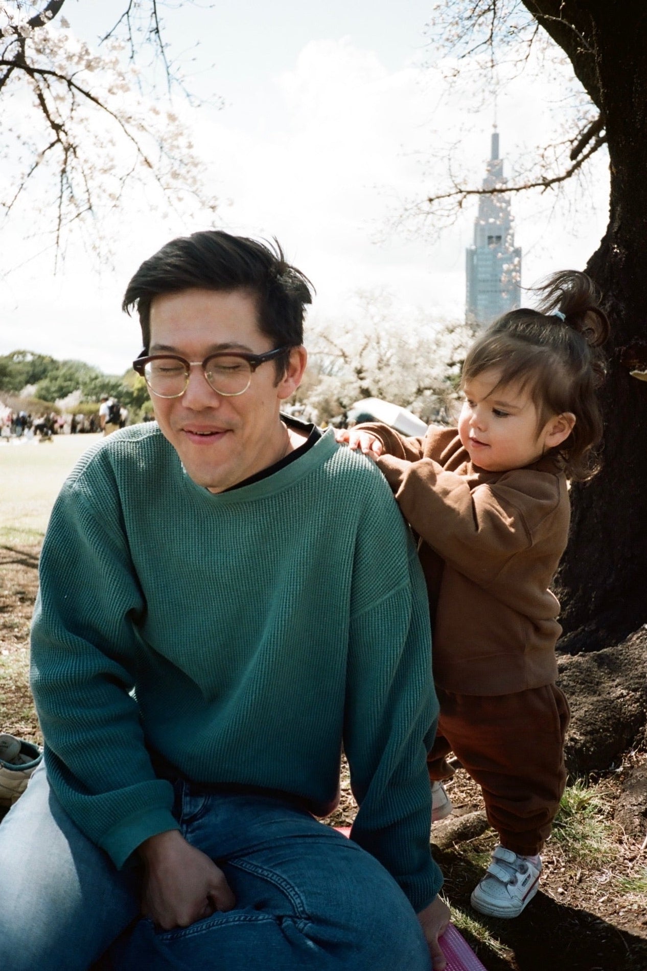 Man holding a child under cherry blossom trees with a cityscape in the background