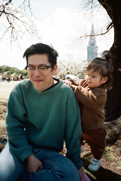 Man holding a child under cherry blossom trees with a cityscape in the background
